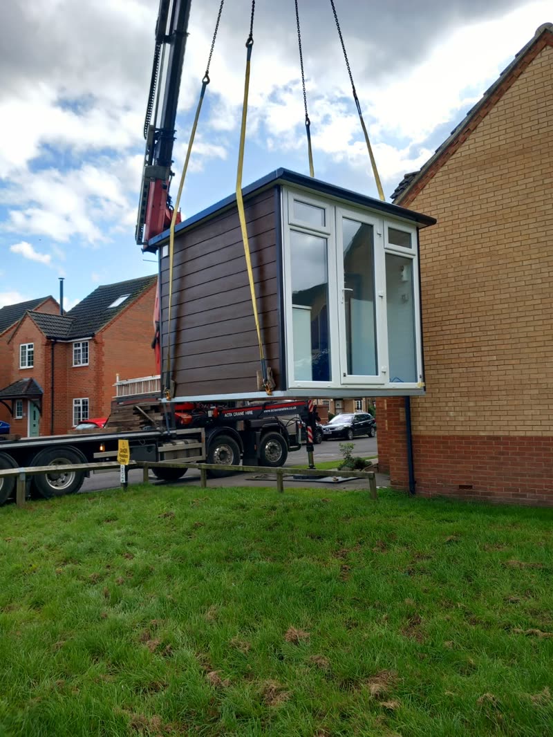 A grey garden room being lifted by a crane onto a lawn beside a brick house
